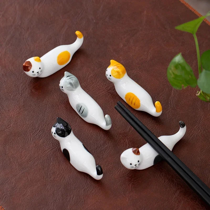 Five ceramic cat-shaped kawaii chopstick holders on a brown surface with a plant in the corner.