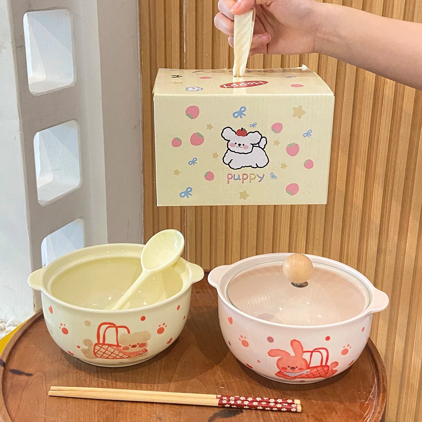 Two children's kawaii ramen bowls with a spoon and chopsticks on a wooden surface, with a branded box in the background.