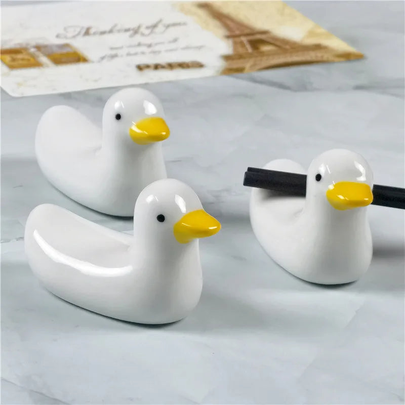 Three white duck-shaped kawaii chopstick holders with yellow beaks on a light gray surface.