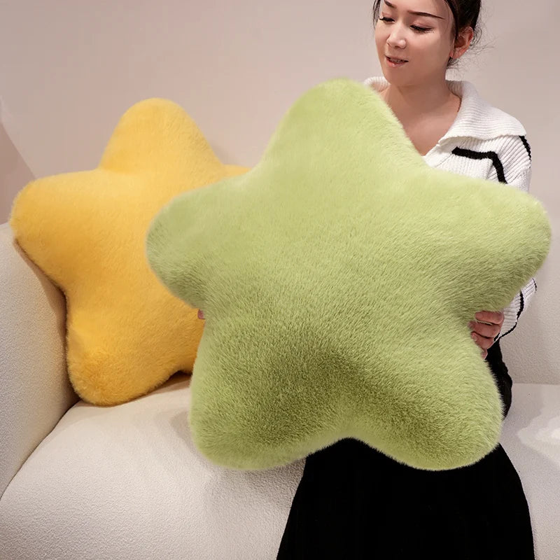 A woman sits on a white sofa holding a large plush star-shaped pillow in light green, with another yellow star pillow resting behind her.