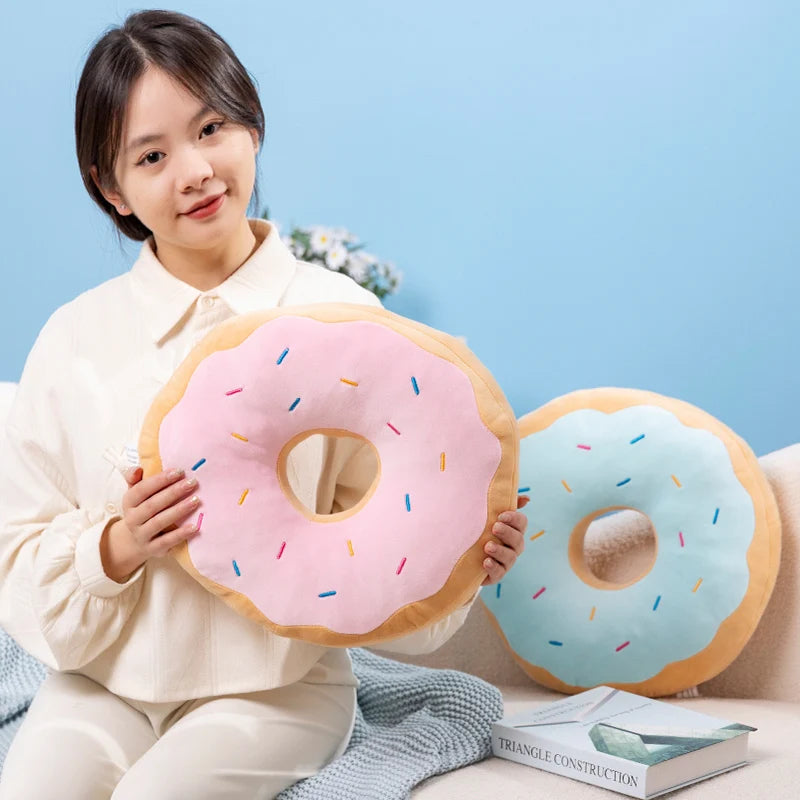 Person holding a donut-shaped kawaii pillow with pink icing and sprinkles against a light blue background.