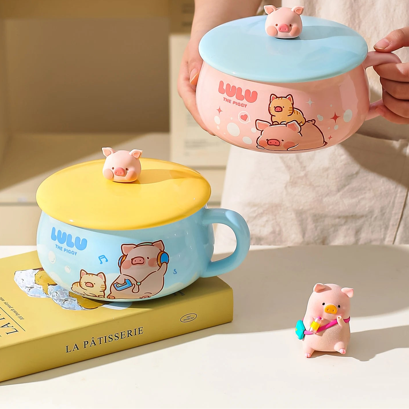Two cartoon-themed ceramic kawaii ramen bowls with pig designs on a surface, one pink and one yellow, with a person holding the pink cup.