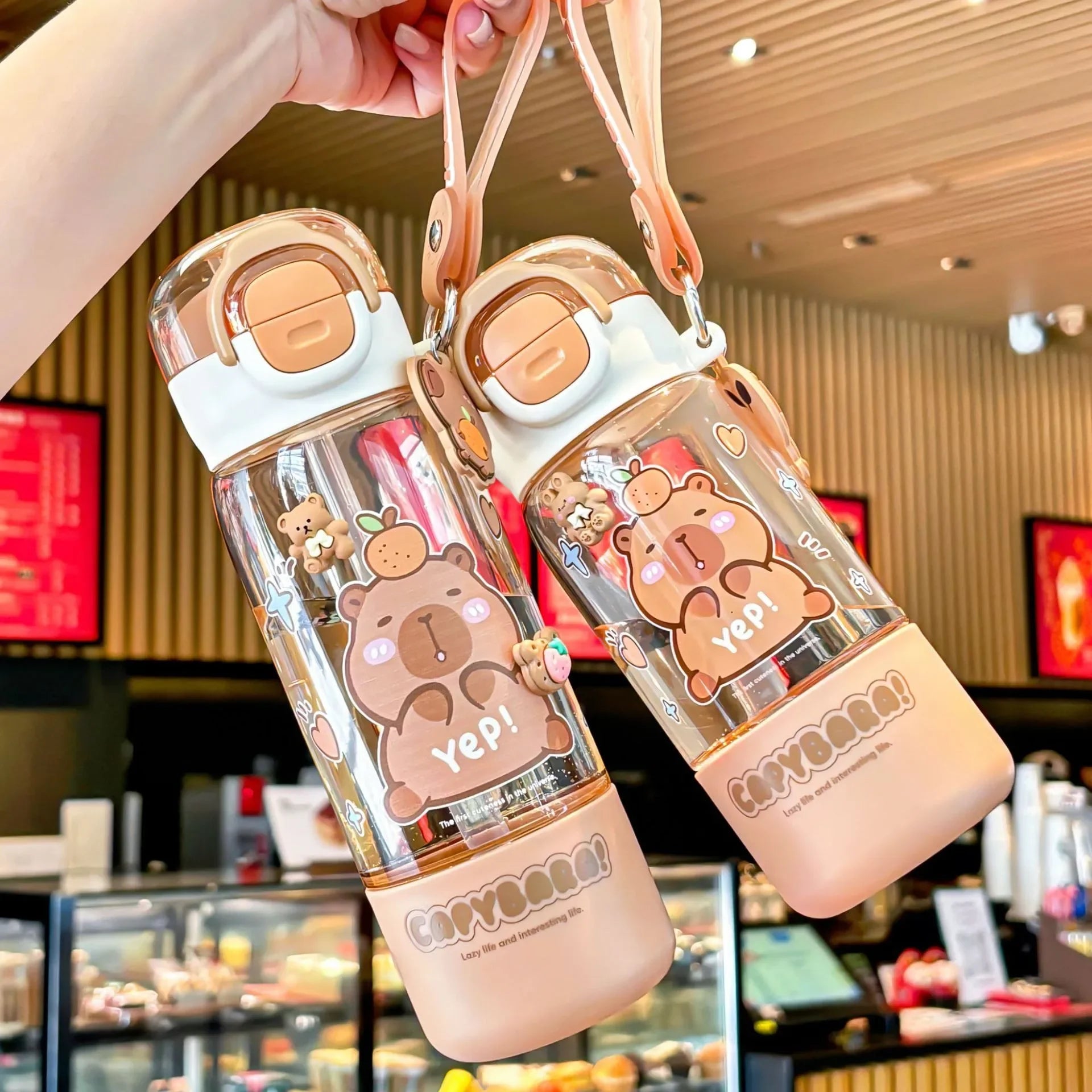 Two cartoon-themed kawaii water bottles held by a hand in an indoor setting.
