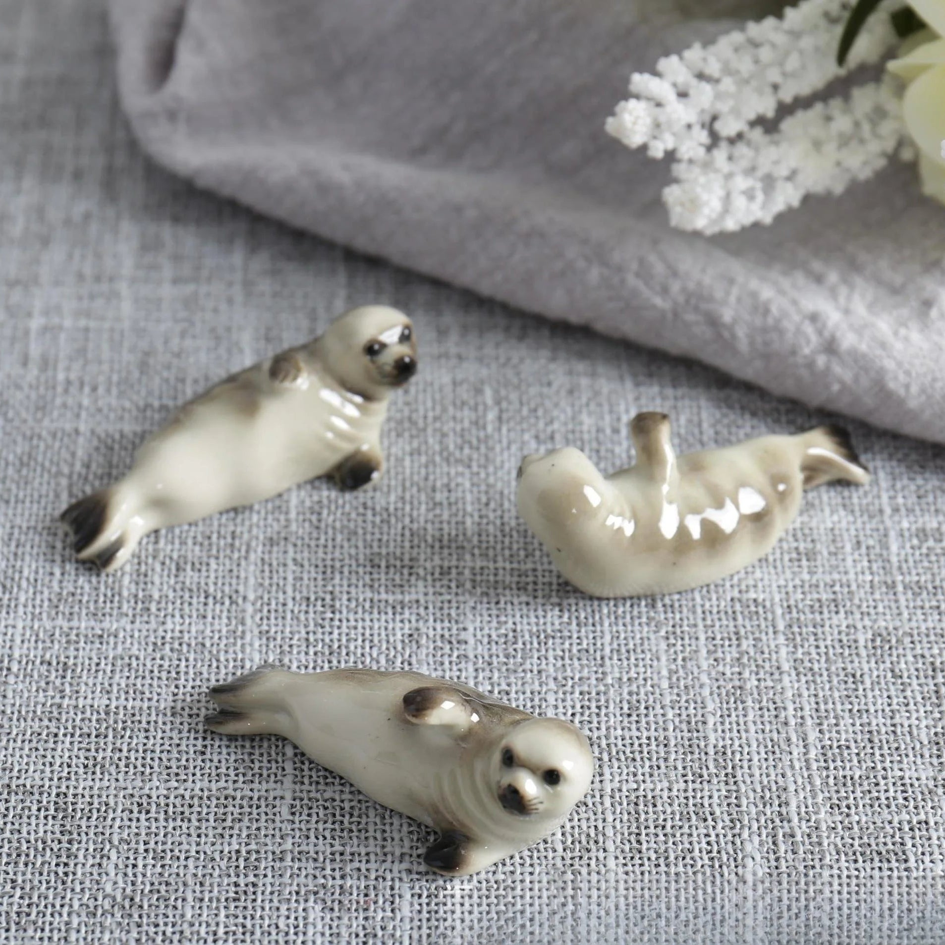 Three ceramic seal kawaii chopstick holders on a textured fabric surface with flowers in the background.