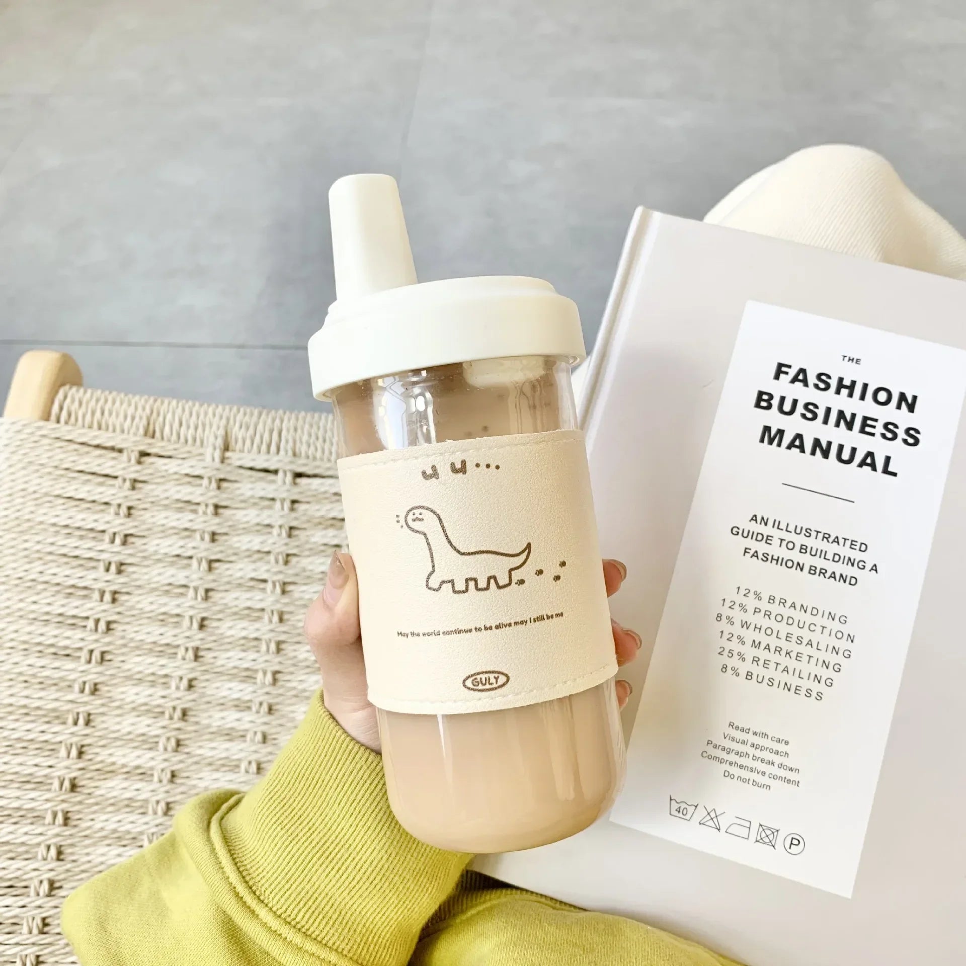 Person holding a kawaii water bottle with a dinosaur label next to a book titled 'The Fashion Business Manual'.
