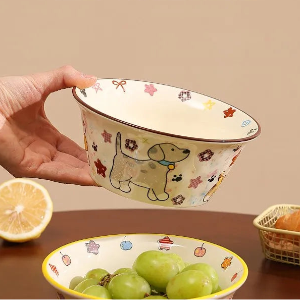 Hand holding a decorative ceramic kawaii ramen bowl with a dog design, filled with green grapes on a wooden surface.