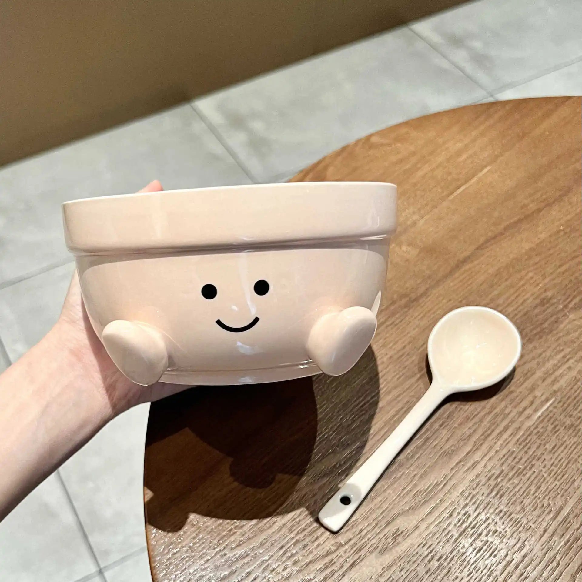 Cute ceramic kawaii bowl with a smiley face design held by a hand on a wooden surface with a wooden spoon.