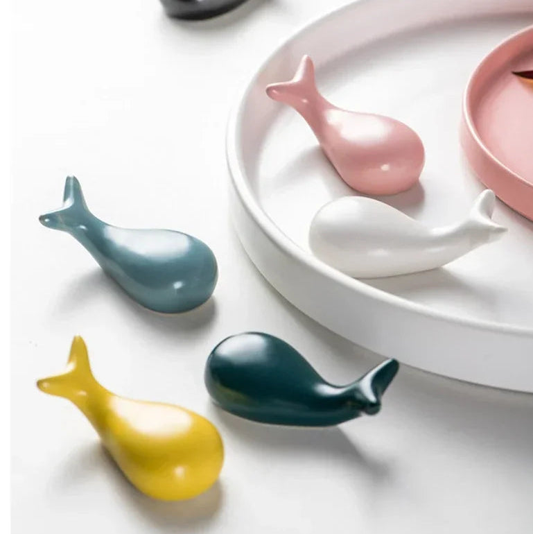 Colorful whale-shaped ceramic kawaii chopstick holders on a white surface with pink and white plates in the background.
