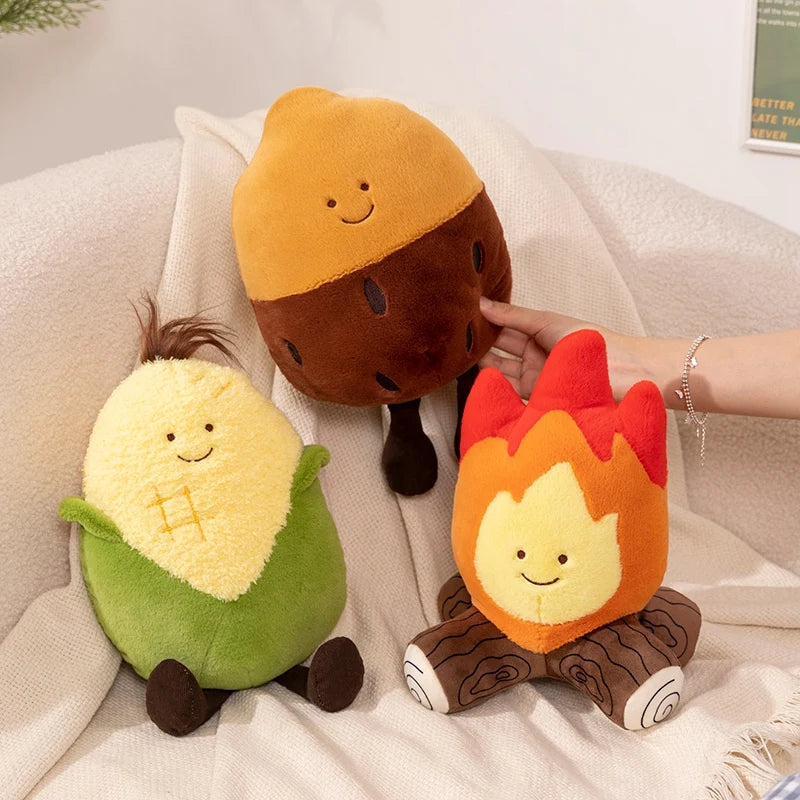 Three cute plush toys shaped like an acorn, corn, and campfire on a soft surface.