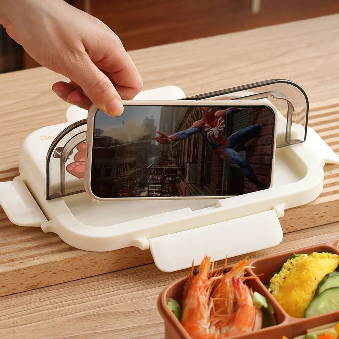 Phone holder on a wooden table with a phone, next to a kawaii bento box with food.