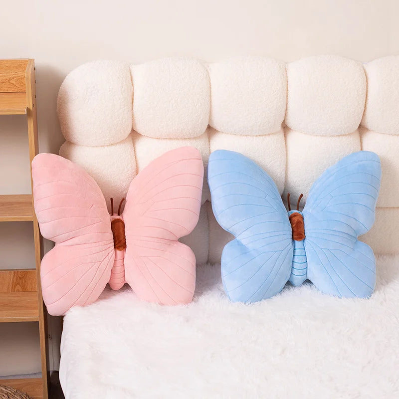 Two butterfly-shaped kawaii cushions, one pink and one blue, on a bed with a white headboard.