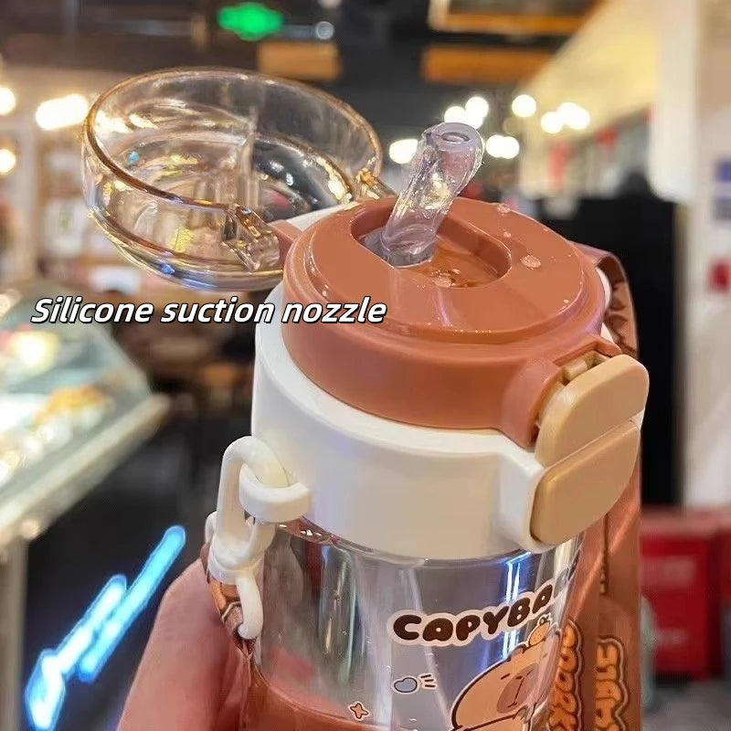 Clear kawaii tumbler with brown lid and silicone suction nozzle held by a hand, blurred indoor background.