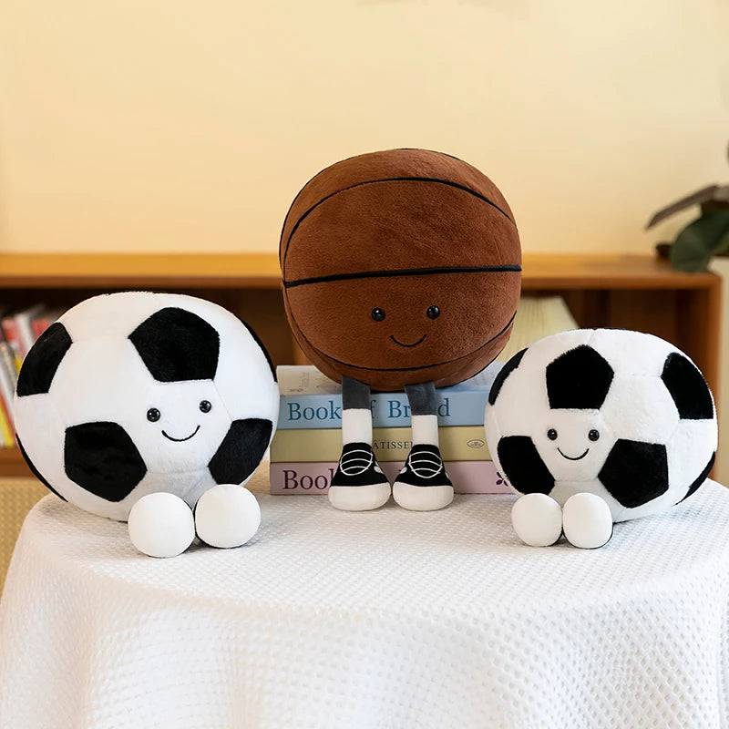 Two kawaii plush soccer balls and a plush basketball on a surface with a neutral background