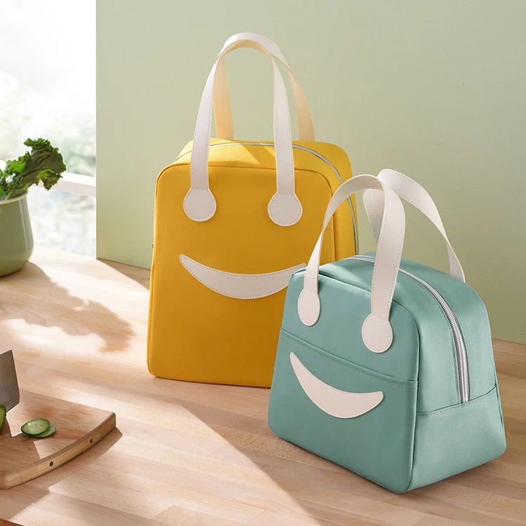 Two smiley face kawaii tote lunch bags, one yellow and one blue, on a wooden surface with a light green wall in the background.