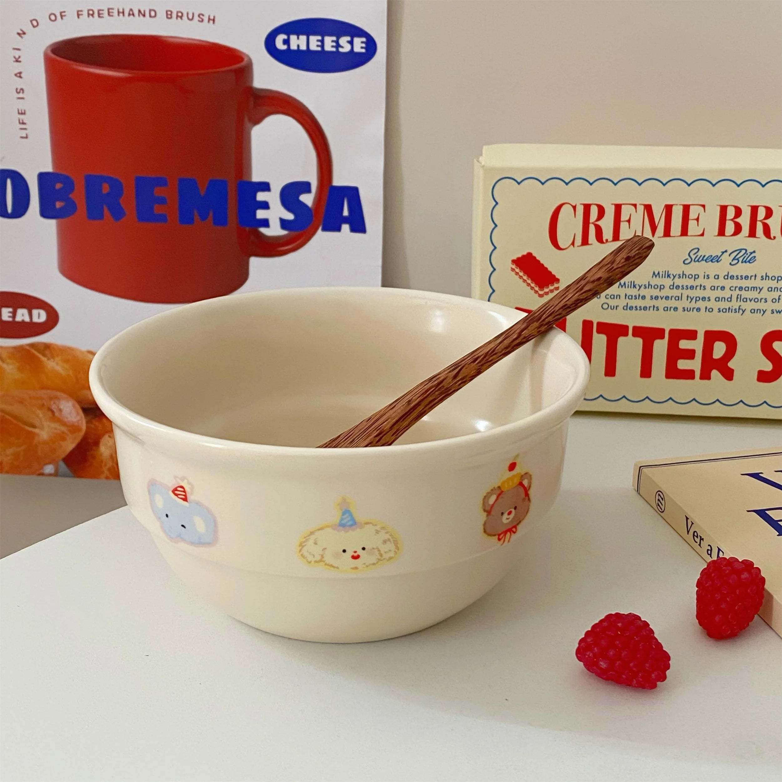 Beige kawaii ramen bowl with cartoon designs, wooden spoon, and red raspberries on a white surface with vintage-style product boxes in the background.