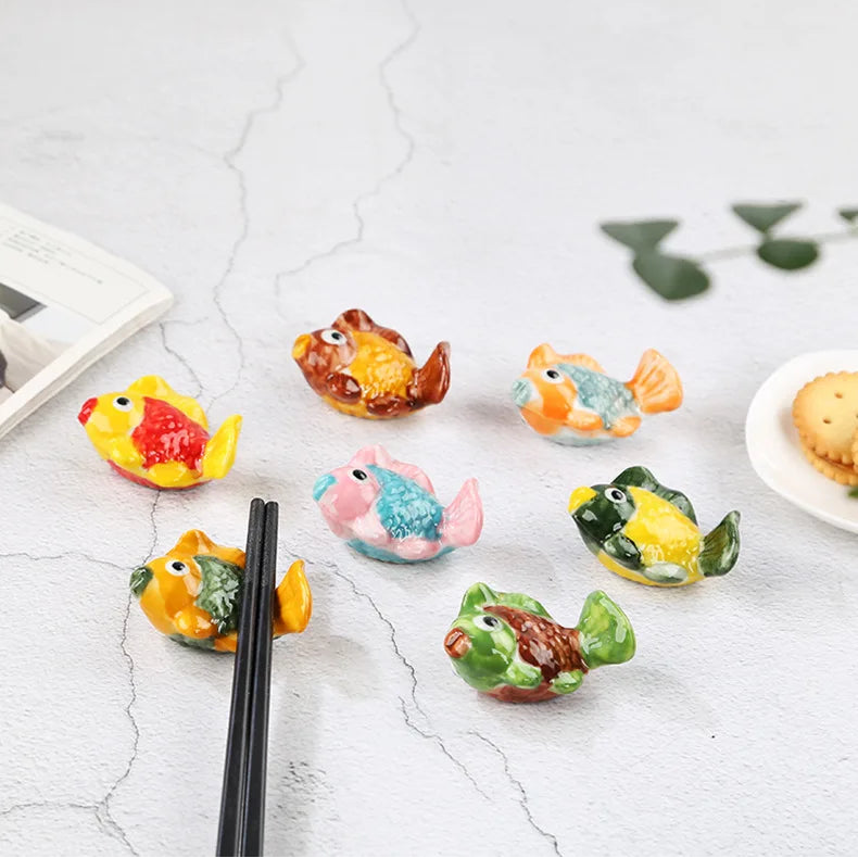 Colorful fish-shaped ceramic kawaii chopsticks holders on a white surface with chopsticks and a plate in the background.