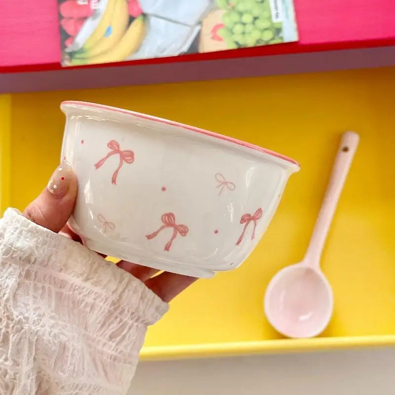 A hand holding a white ceramic bowl decorated with pink bow and dot patterns, with a matching pink spoon resting in a yellow tray behind it.