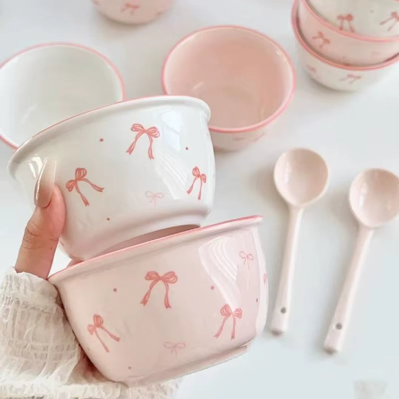 A hand holds two small ceramic bowls decorated with pink bow and dot patterns on a white background, with matching spoons and additional bowls around them.
