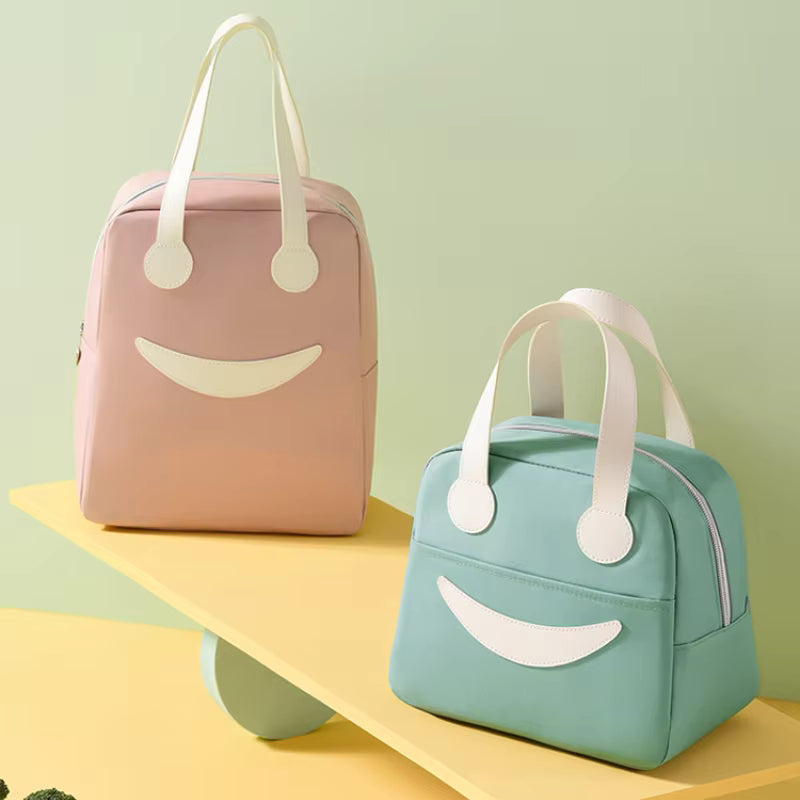 Two cute smiley-face insulated lunch bags displayed on a yellow platform against a pastel green background. One bag is light pink, and the other is light mint green.