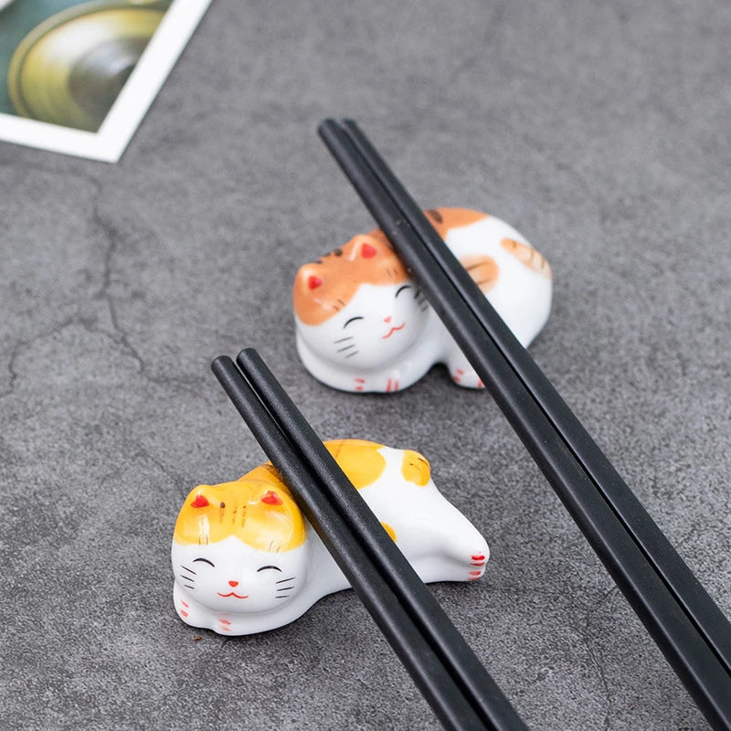 Two kawaii cat-shaped ceramic chopstick rests on a gray surface with black chopsticks.