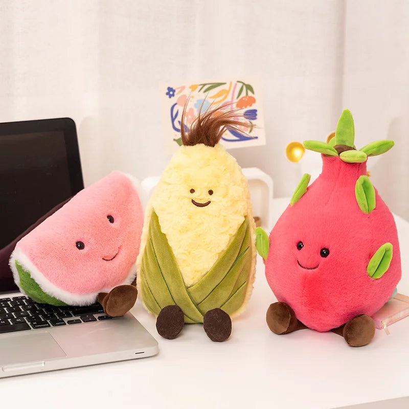 Three cute plush fruit toys on a white desk: a pink watermelon slice plush leaning on a laptop, a yellow corn-on-the-cob plush wrapped in green leaves, and a pink strawberry plush with green leaves and brown feet. A small card is tucked behind the corn plush.