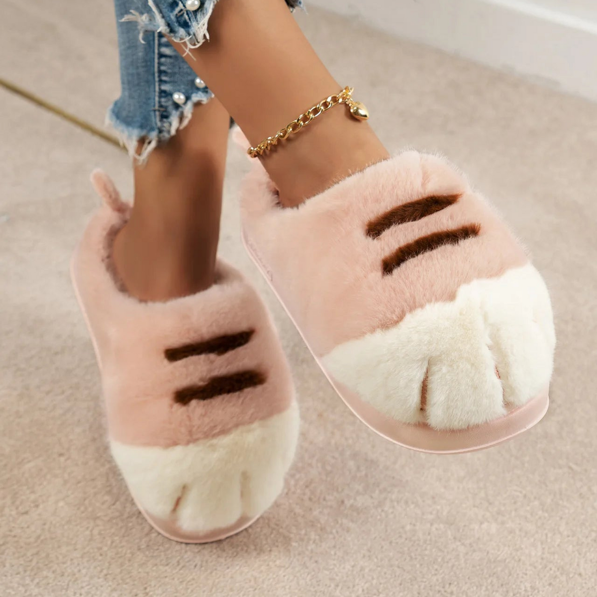A person wearing fluffy cat-themed slippers with pink and white faux fur and dark brown claw details, paired with frayed blue jeans and a gold anklet, standing on a beige carpet.
