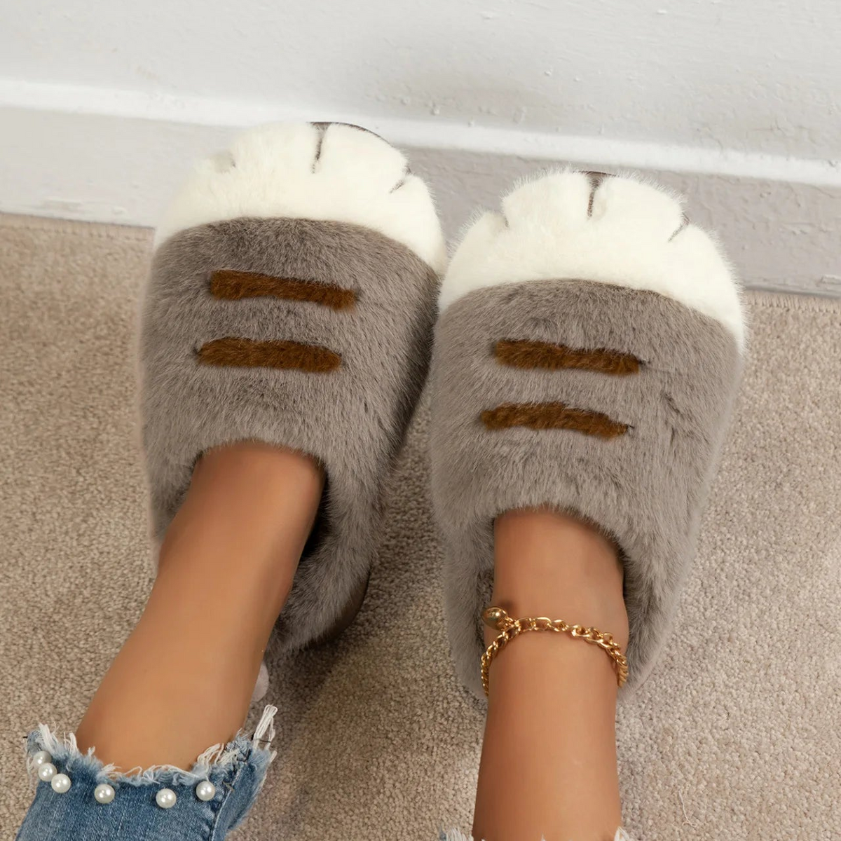 A person wearing plush cat-look slippers with gray bodies, white paw-clip tops, and brown stripe details, standing on a beige carpet near a white baseboard.