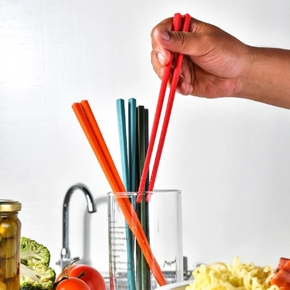 Hand holding red kawaii chopsticks with a glass containing blue and black straws, vegetables, and pasta in the background.