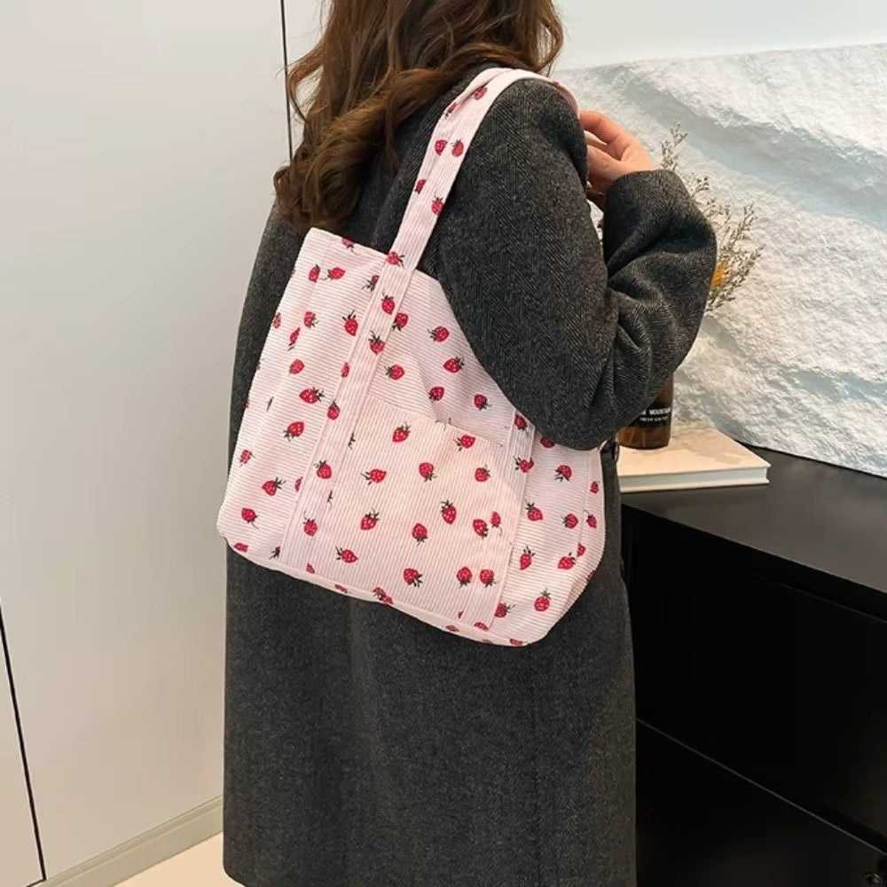 A woman wearing a gray coat carries a large pink tote bag with a red strawberry print in an indoor setting.