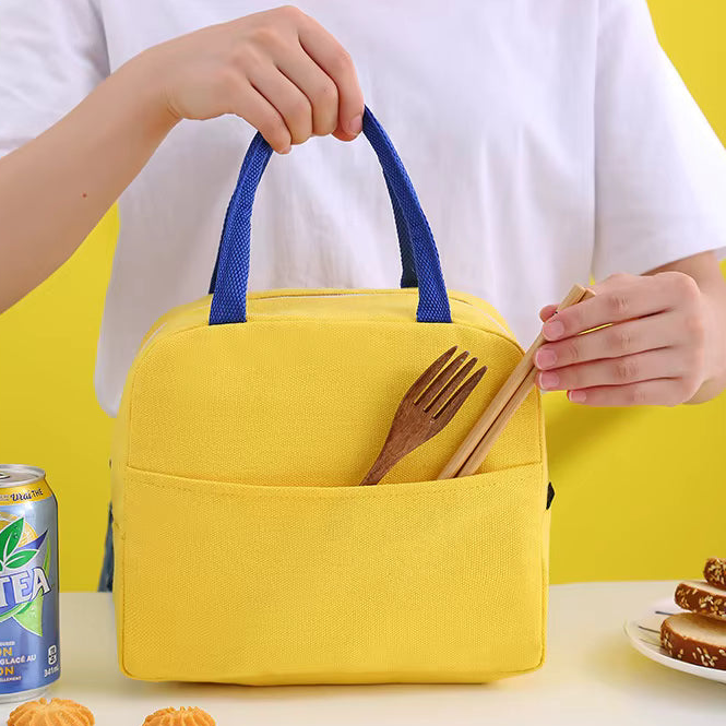 A bright yellow insulated lunch bag with blue handles is held by a person wearing a white shirt. The bag has a front pocket containing wooden chopsticks and a wooden fork, set against a bright yellow background with a soda can and a plate of food on a table below.
