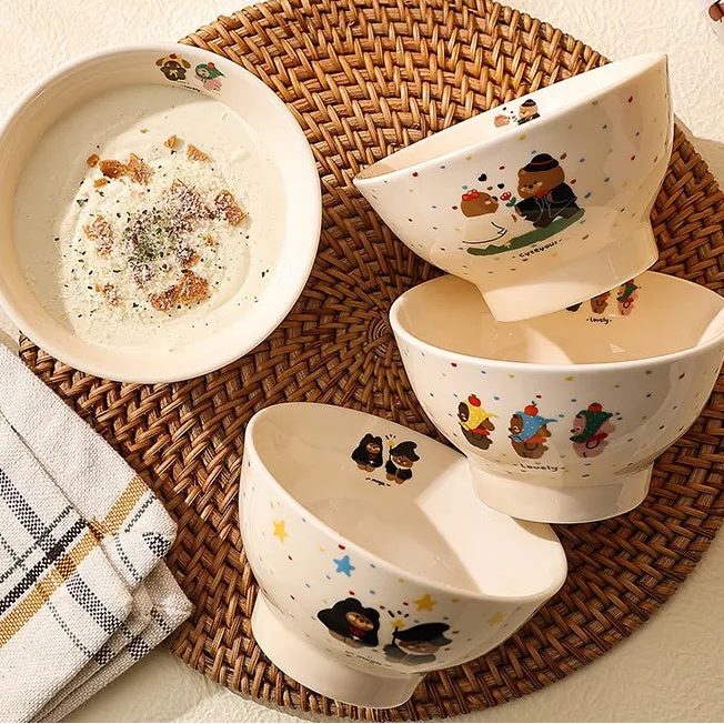 Set of cream-colored kawaii ramen bowls with bear designs on a woven mat with chopsticks and fruit.