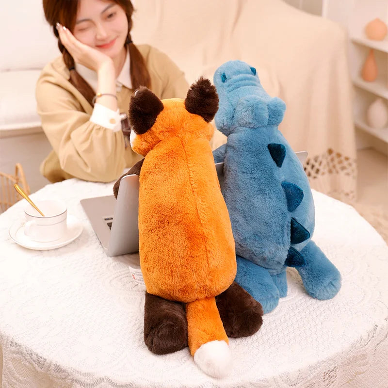 Two kawaii plush toys, one orange and one blue, sitting on a table with a person using a laptop in the background.