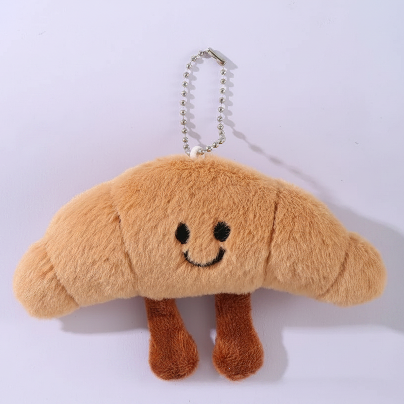 A plush croissant keychain charm with a smiling face and a silver ball chain, shown on a light background with measurement lines and Chinese text indicating dimensions.