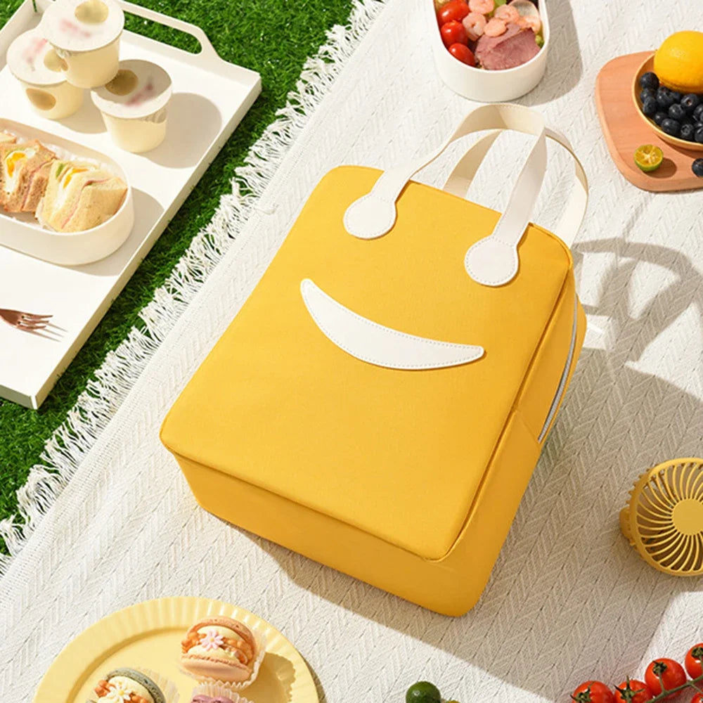Yellow kawaii lunch bag with smiley face design on a picnic setup with food and a fan.