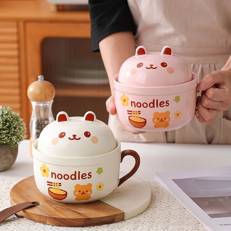 Two cute ceramic kawaii ramen bowls with animal faces and 'noodles' text, one pink and one white, on a wooden surface.