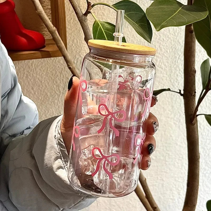 Clear glass kawaii tumbler with pink bows held by a person, with a plant in the background.