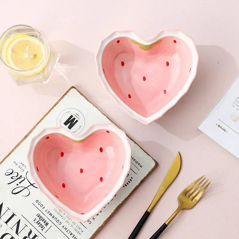 Heart-shaped pink ceramic kawaii bowls with watermelon pattern on a light pink surface with a candle and cutlery.