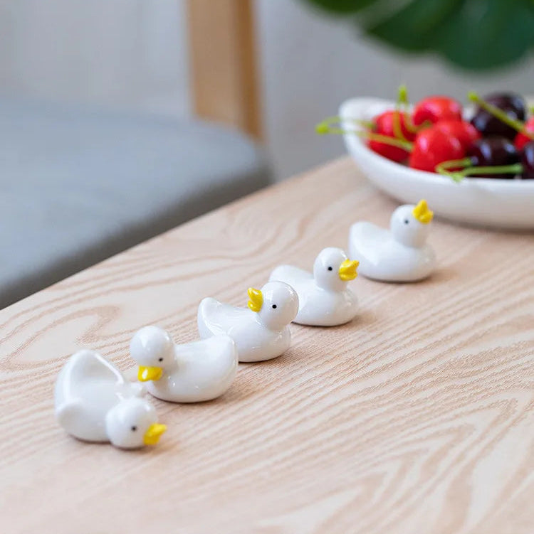 Small white ducks with yellow beaks kawaii chopstick holders on a wooden surface, with a bowl of cherries in the background.