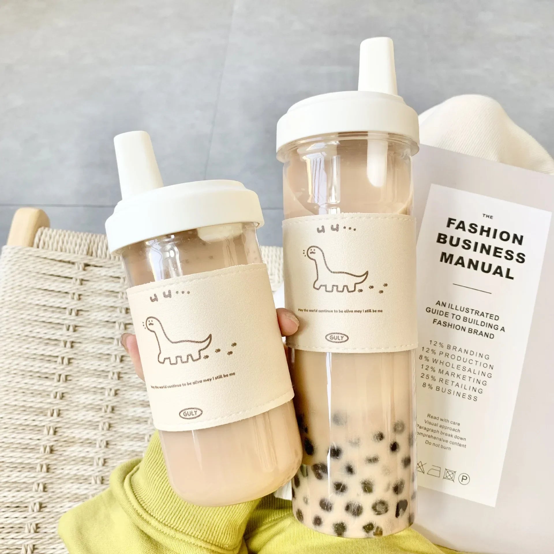 Two bubble tea kawaii bottles with straws and a book titled 'Fashion Business Manual' on a light gray background.