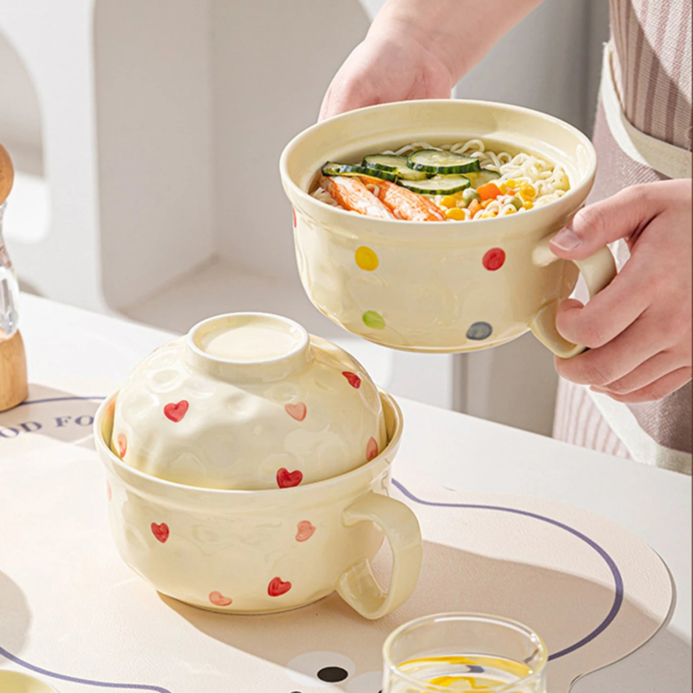 Person holding a kawaii ramen bowl of soup with a heart-patterned lid on a table.