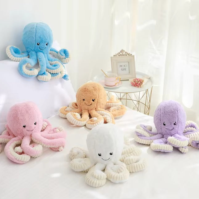 Five plush octopus toys in pastel colors are arranged on a white bed with soft curtains and a small side table in the background. The octopuses have soft fuzzy bodies and ribbed tentacles, each with embroidered smiling faces.