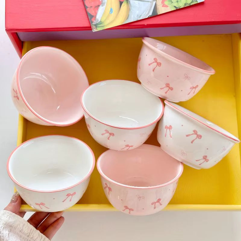 Six pastel pink and white ceramic bowls with a pink bow pattern, arranged inside a yellow tray with a red lid, held by a hand at the bottom left.