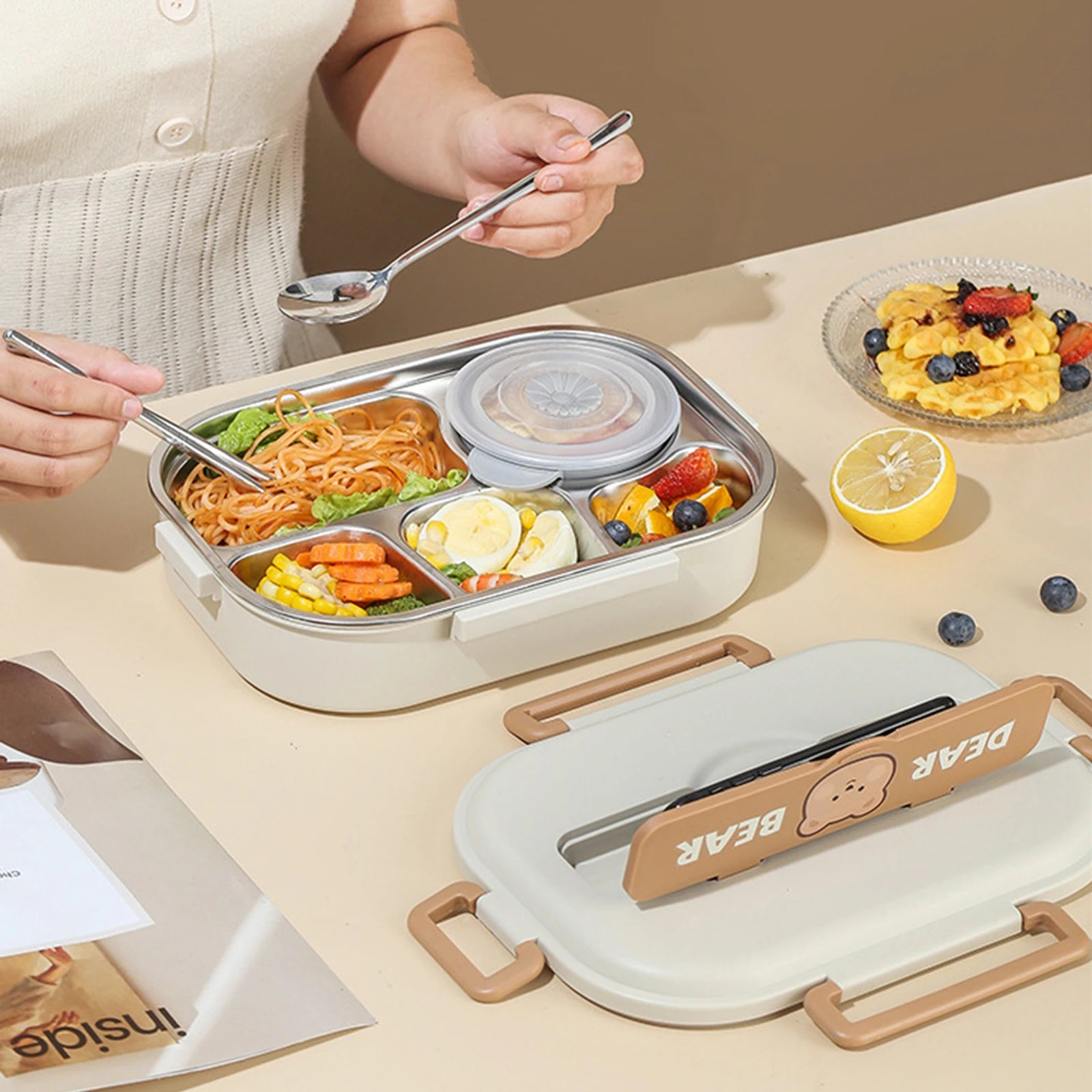 Person eating from a compartmentalized kawaii lunch box with a side of fruit on a table.