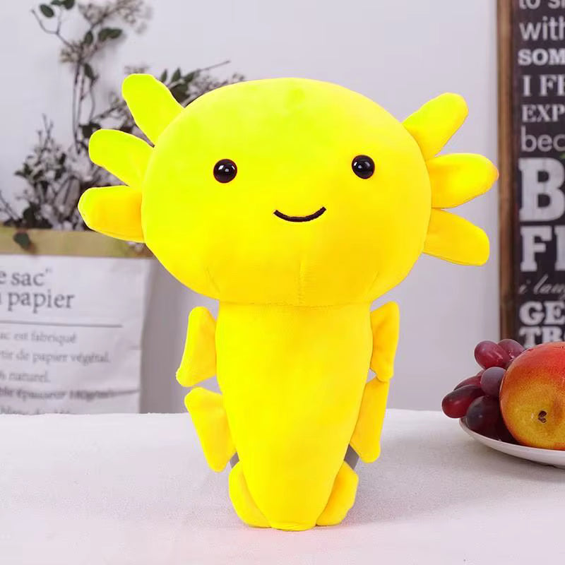 A bright yellow plush axolotl toy with embroidered black eyes and a smile, standing on a white tabletop with decorative background elements including a fruit plate and paper bags.