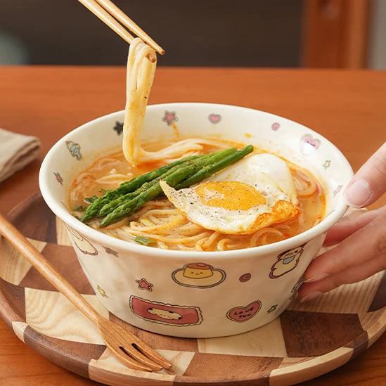 Person holding a kawaii ramen bowl of ramen with chopsticks on a wooden table.