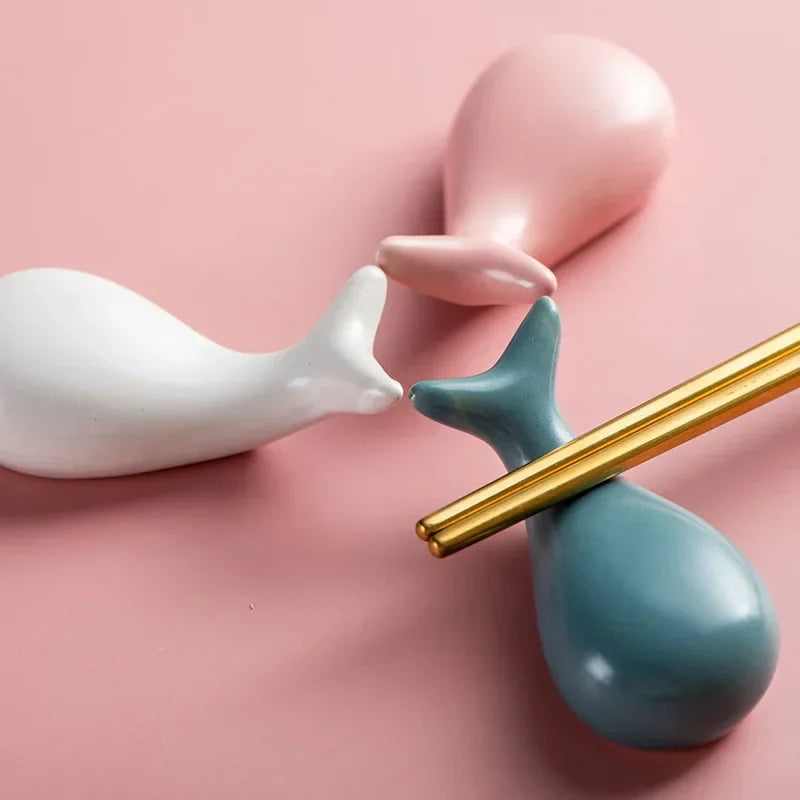 Two ceramic kawaii chopstick holders, one white and one blue, with a gold object between them on a pink background.