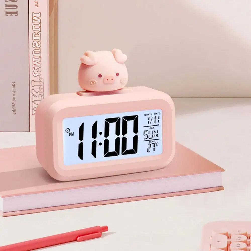 Pink pig-shaped kawaii alarm clock on a light pink surface with a pen and notebook.