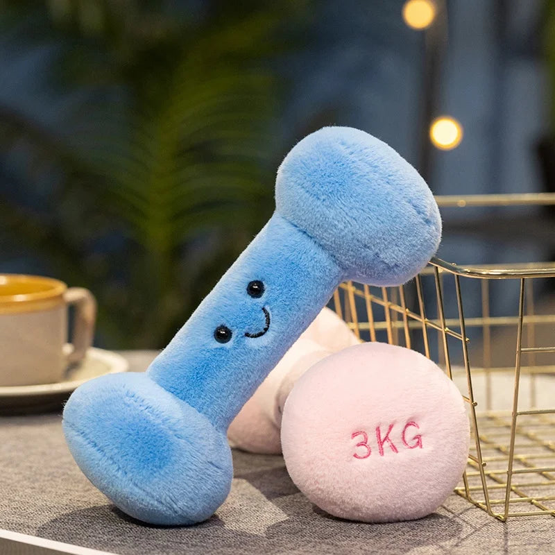 Blue kawaii plush dumbbell with a smiling face next to a pink dumbbell labeled '3KG' on a blurred background.