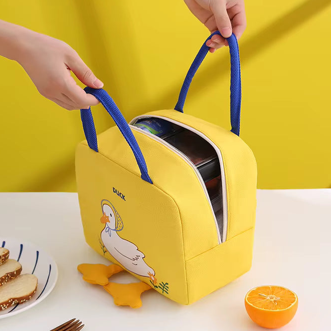 A yellow soft-sided lunch bag with blue handles, shown on a white table with hands holding the handles. The bag has a cartoon duck illustration and a partially open top zipper revealing packaged items inside.