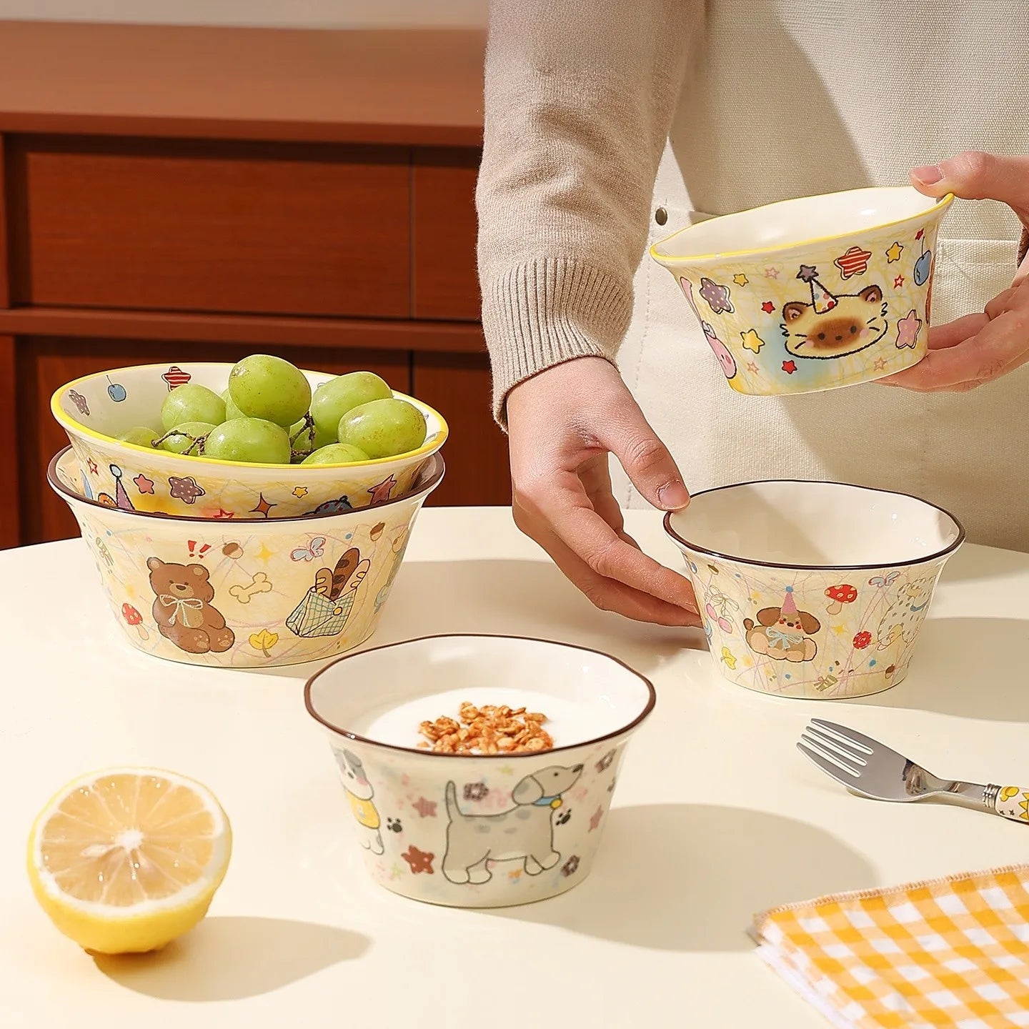 Person holding a kawaii ramen bowl with cartoon designs on a table with other bowls and a lemon.